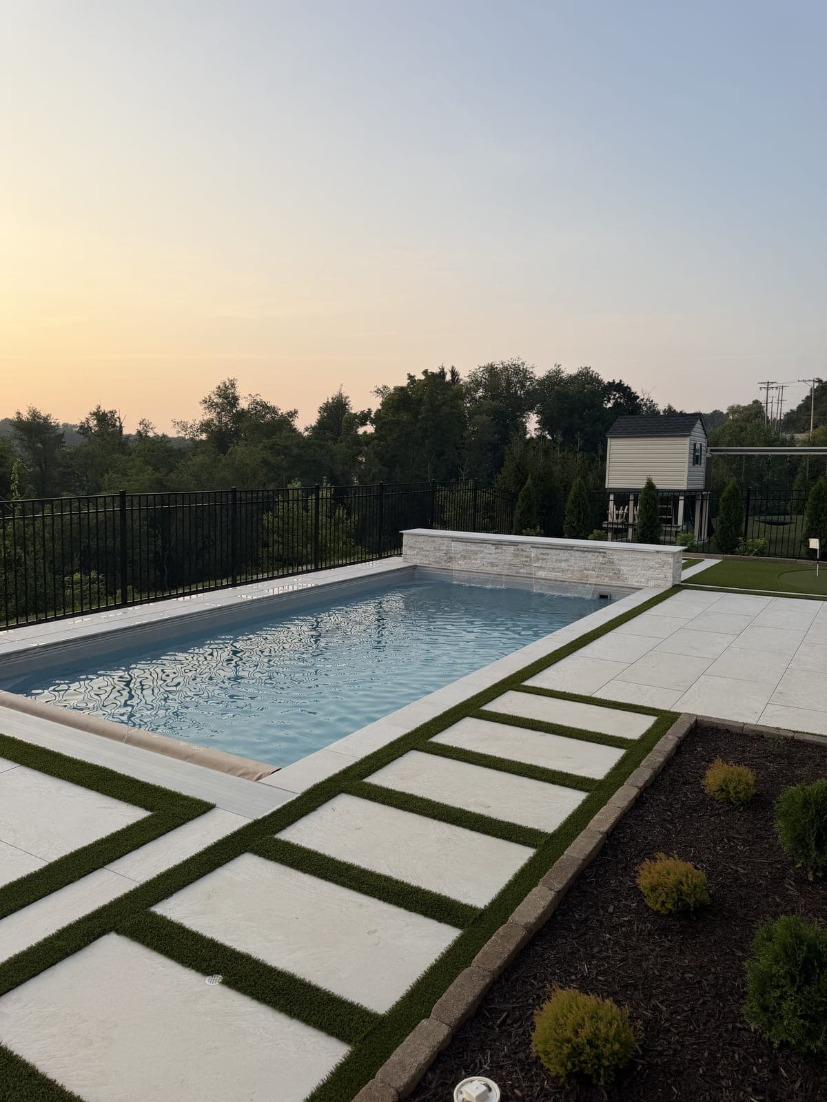 Pool with stacked-stone waterfall wall and checkerboard paver-turf deck at sunset