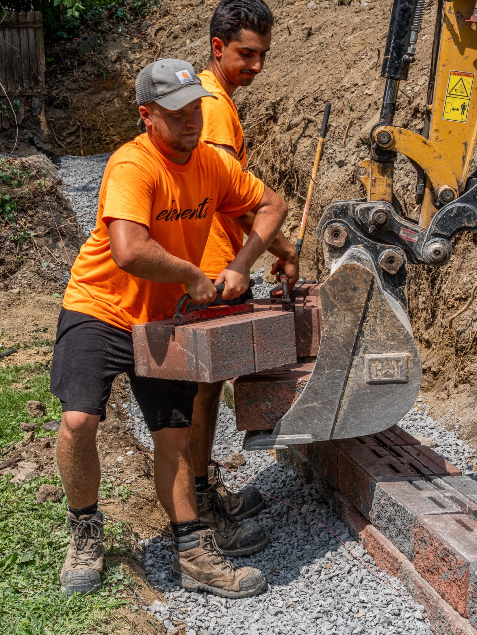 Elements crew setting a wall block on the gravel base course beside an excavator bucket